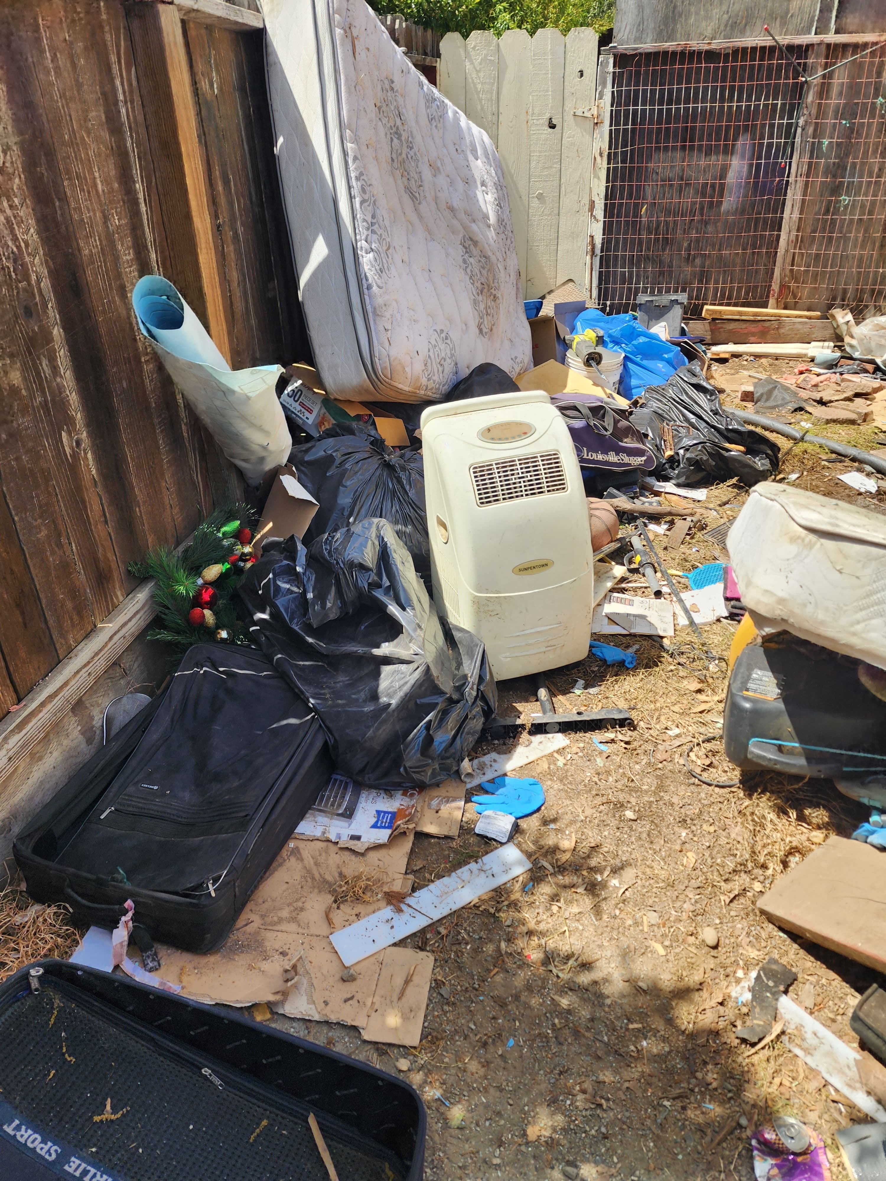 Cluttered alleyway with discarded mattresses, trash, and debris under a wooden fence.